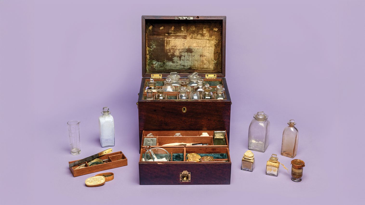 Brown wooden chest displayed open with its contents arranged next to it, including small glass vials and jars, against a light purple background